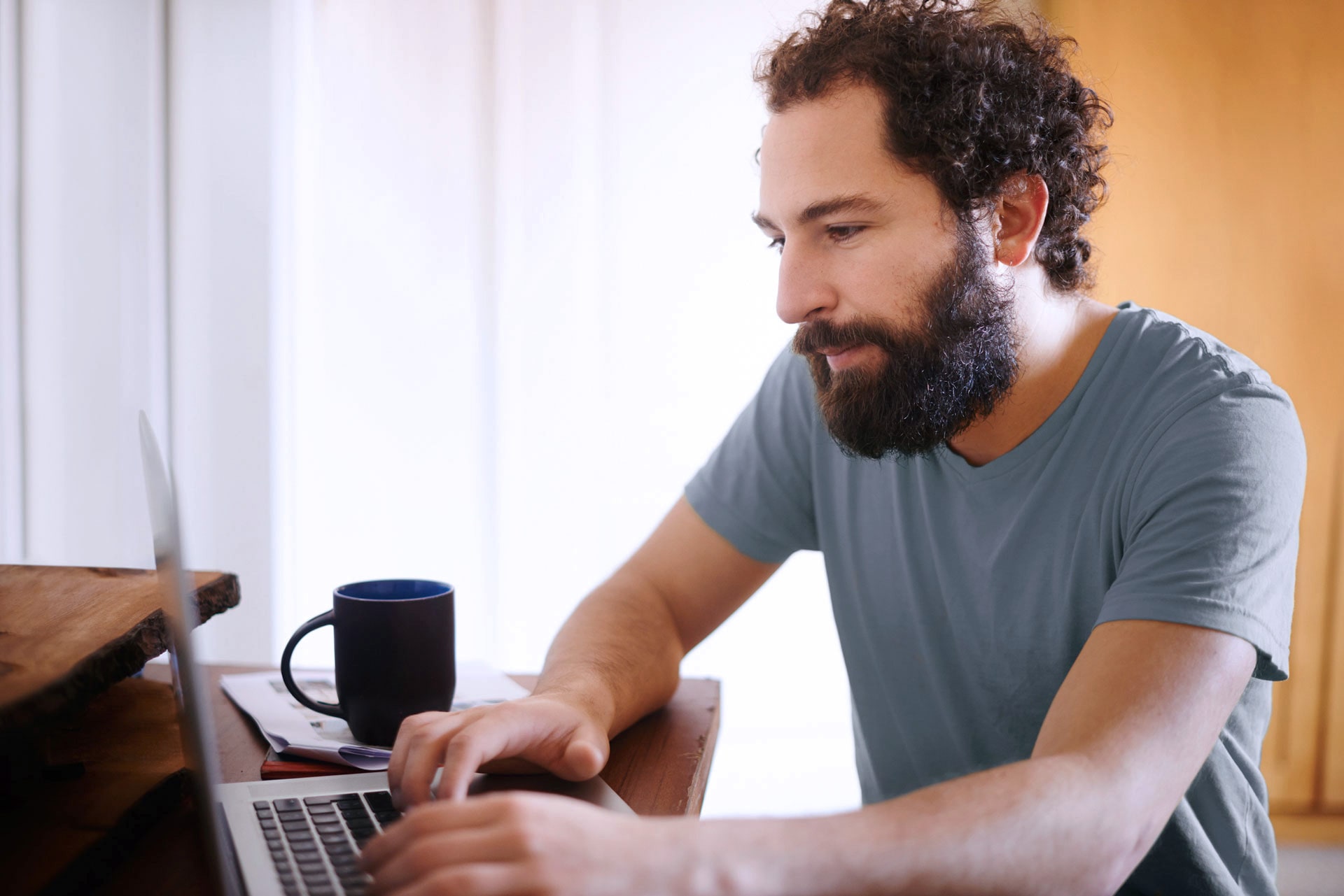 Ein junger Mann mit Bart sitzt tippend vor einem Computer. Neben ihm befindet sich eine Kaffeetasse.